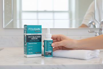 Hand holding FOLIGAIN DHT serum beside its box and a folded towel on a bathroom counter.