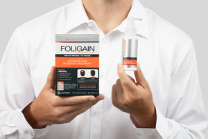 Man holding FOLIGAIN 5% minoxidil and 5% TRIOXIDIL topical treatment box and spray bottle.