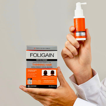 Hands holding FOLIGAIN men’s 10% TRIOXIDIL topical treatment box and spray bottle.