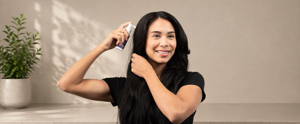 Woman parting her hair and holding a FOLIGAIN TRIOXIDIL spray applicator near her scalp.